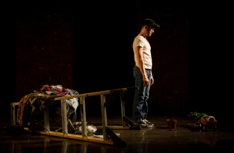 On a warmly lit stage, a dancer stands upright, chin slightly dropped. Behind them is a horizontal ladder draped with articles of clothing. Photo by Ian W. Douglas.