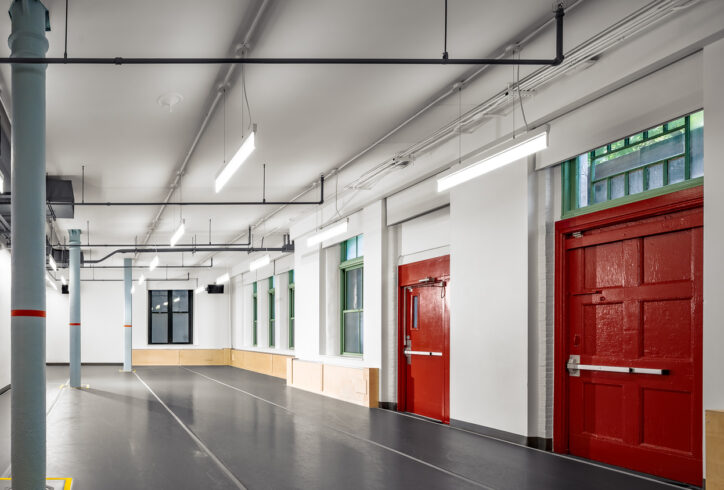 A dance studio with a grey marley floor, windows, and red doors. There are three grey columns distributed through two thirds of the space. Photo by Alexander Severin .