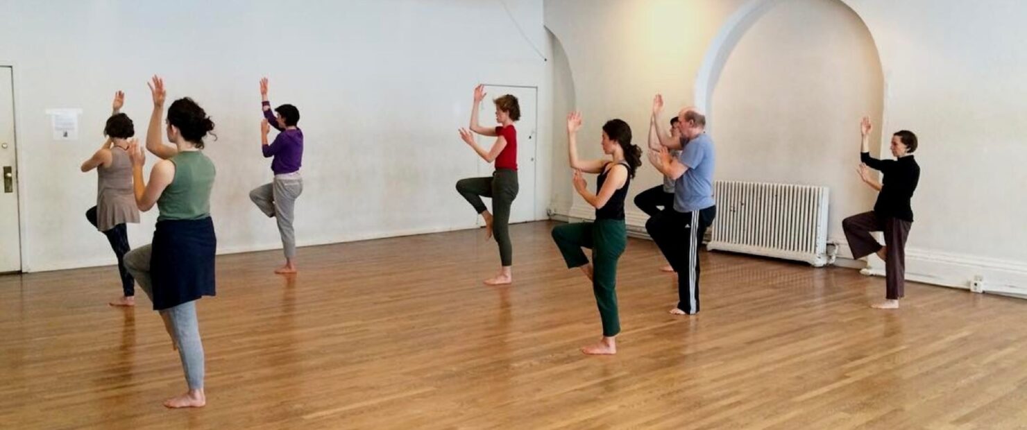 A group of dancers in a light space lift their right leg in unison, bringing their right foot to their left knee. Lifting their right arm up at a right angle with the left arm just below. 