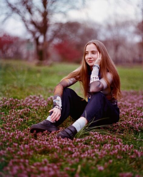 Photo of Tess, a white woman with long dark blonde hair, sits in a grassy field with pink flowers looking out with her head resting on one hand. Photo by Gus Aronson.