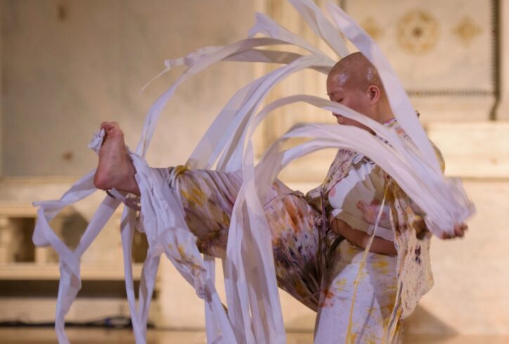 A dancer performing at Movement Research at the Judson Church. One leg is in the air while swirling strips of fabric flow around them. Photo by Ian Douglas.