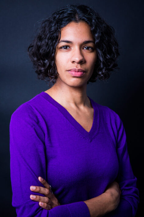 Portrait of Marýa Wethers. She has black curly hair and a bright purple top. She looks into the camera with a neutral expression. Her arms are crossed in front of her. Photo by Scott Shaw.