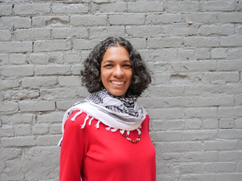 Portrait of Marýa Wethers. She has black curly hair and wears a long sleeve red top and a keffiyeh. She looks into the camera smiling brightly. Photo by Katherine Paola De La Cruz