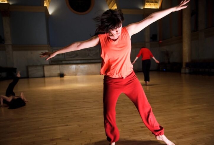 A dancer in red pants and an orange top has their arms out in a T, one leg is hovering over the ground, and they look down at the Judson Church.