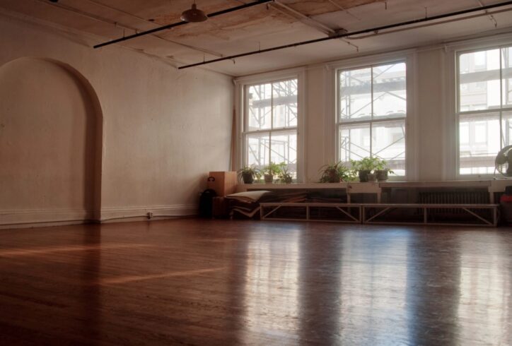 Empty dance hall, with 3 large windows, reflected in the wooden floor. There are plants in the window sills.