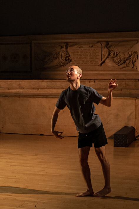 Glenn Potter-Takata dances for Movement Research at the Judson Church. He is stands tilting forwards with his arms away from his body curving and contorting in different directions. Photo by Rachel Keane.