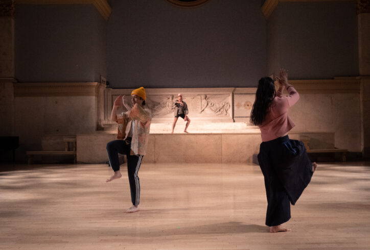 Three dancers performing at Judson Memorial Church. MR Artist-in-Residence, Glenn Potter Takata is in a squat position in the altar area. Two dancers are at level floor in separate corners and facing opposite directions. Their hands at face level one knee elevated at hip level. Photo by Iki Nakagawa.