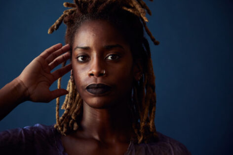 Angie Pittman poses for a headshot. Her hand reaches towards her blond and brown locs as she looks into the camera with a neutral expression. Photo by Whitney Browne.