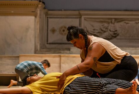 Two dancers at level floor during Sara Shelton Mann's class. One is lying on their back with arms and legs extended out while the other is kneeling next to them with hands gently touching the pelvis of the dancer at rest. Behind them another dancer can be seen doing the same holding gesture. The Judson Memorial Church altar serves as background. Photo by Rachel Keane.