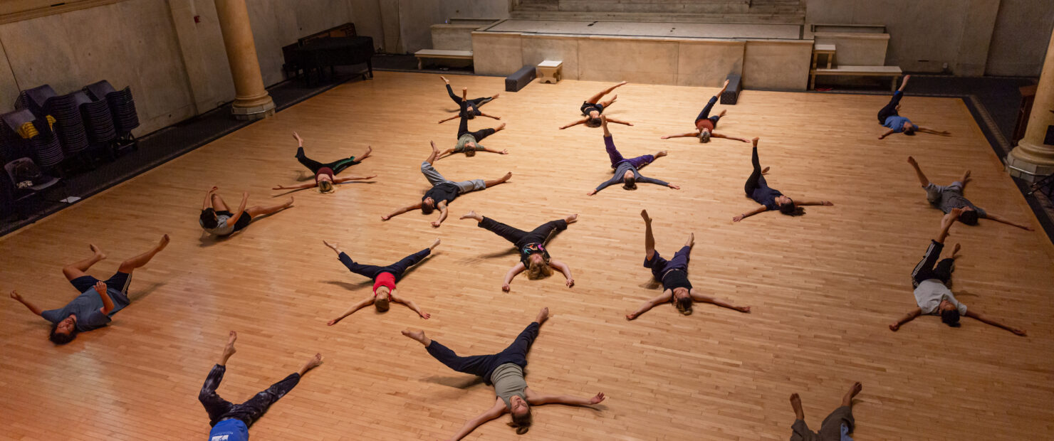 Image taken from above. MELT Summer 2023 Workshop taught by Joanna Kotze at Judson Memorial Church. Dancers lie on their backs with arms spread out. One leg is also extended out as the other seems to be in motion bending at the knee and floating of the ground. Photo by Rachel Keane.