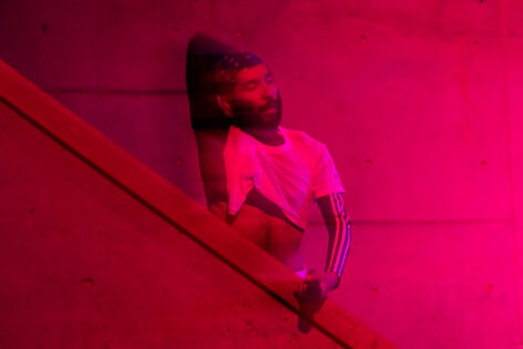 Estrellx is standing on a staircase with their right arm-elbow over their head, eyes closed, while their left hand gently holds a mic; the lighting is a soft fuschia that illuminates Estrellx's white mesh crop top and dark green Adidas long sleeve shirt. Photo by Elyse Mertz.