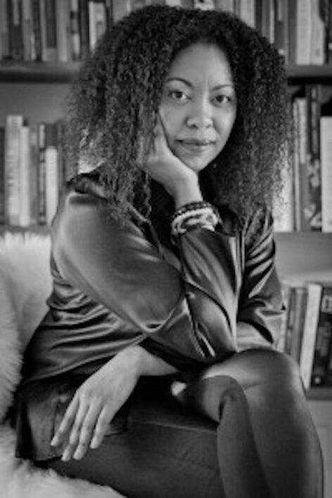Black and white portrait of Adrienne Edwards. She looks into the camera with a neutral expression. Her curly hair frames her face and she rests her head on her hand. She is seated leaning forwards slightly. The other hand rests across her lap. Photo by Whitney Browne.