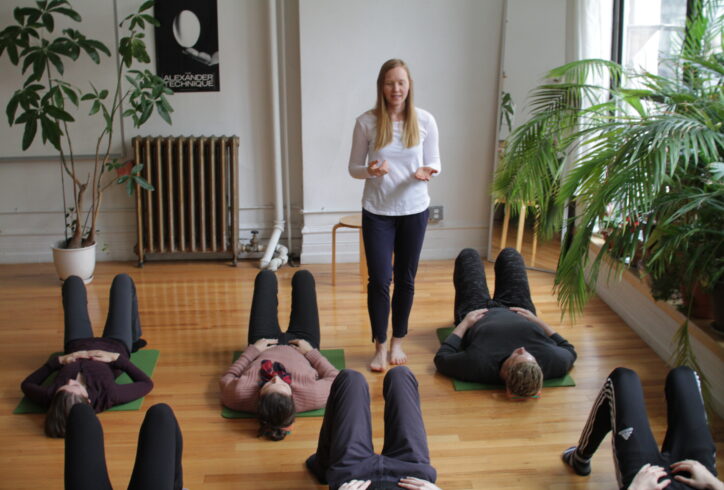 Photo shows Alice teaching in a room with plants, white walls, windows, and a hard wood floor. Students lie in two rows on the floor on their backs. Alice is walking among the students speaking as she teaches.
