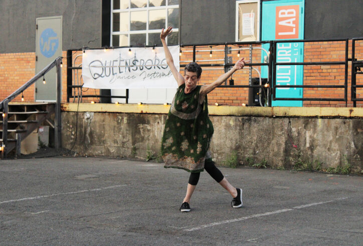 Barbara Mahler dancing in a green dress. Arms outstretched, standing in front of a sign that reads Queensboro Dance Festival, and a loading ledge. Photo courtesy of the artist.