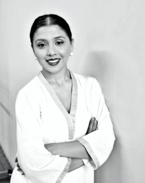 A black and white photo of Barkha Patel, a South Asian woman wearing a white dress. Her hair is parted in center and tied away from her face, her arms are crossed in front of her and she is smiling softly into the camera.Photo courtesy of the artist. Photo courtesy of the artist.