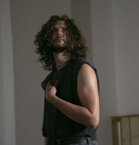 Image of a performer with brown shoulder-length curly hair and a short beard. They are cropped from the hips up and wearing a black tank top. Their upper body is pivoting front and their eyes are looking
quizzically to the upper right diagonal with their right hand pointing to their chest. Photo by Whitney Browne.