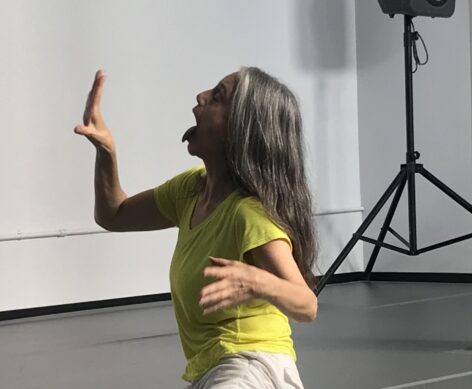 Daria, a woman with light olive skin, wears a yellow T-shirt and white pants. She kneels on the floor facing diagonal left, her wait is on the far knee. Her left hand, palm facing out, is in front of her face, and she sticks her long tongue out. She has long dark and silver hair hanging on her back. Photo courtesy of the artist. 