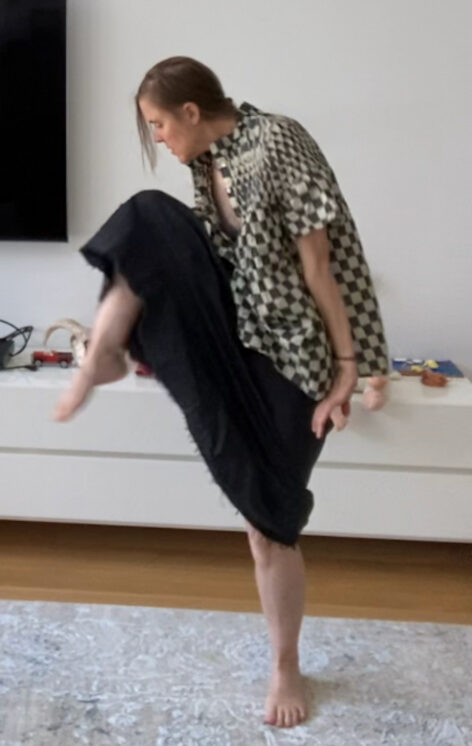 Emily wears a flowly black and white checkered top and a dark skirt. Her right knee is raised higher towards her face. Her left leg is straight on the ground. She appears to be dancing in a living room. Photo courtesy of the artist.