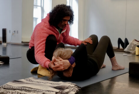 Antonio Ramos offering tactile support to a student during class. The student is on yoga mat on the floor, hands behind their head, feet on the ground, knees bent and the head slightly off the ground. Antonio is holding the student's head with one hand and the other is rested just above the left knee. Antonios curly hair falls on his face. Photo by Harry Shunyao Zhang.