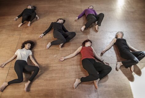Six people engaged in a movement lying on their backs on a floor with both knees bent and tilted to the left.Their arms are relatively close to their torso with palms facing up. Each person has their head turned different degrees to the left in quiet repose. Photo Courtesy of Tasha Taylor.