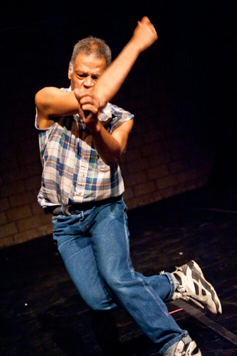 Solo brown-skinned male dancer jumping while wearing a plaid shirt, jeans and high-top sneakers. Photo by Ian Douglas.