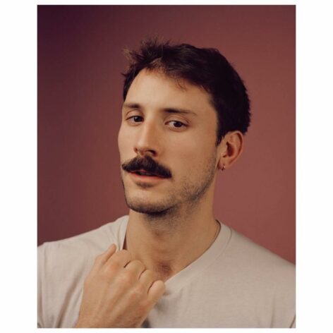 John Arthur Peetz poses for the camera. He wears a white T-shirt and holds the collar with his hand. He has dark hair and a full mustache. The background is pink. Photo by Ryan Pfluger.