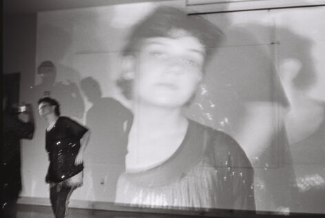A black and white photo of a person with dark curly hair posing for a photographer. Behind them is a projection showing a large close up of their face. Photo by Aisha Tida.