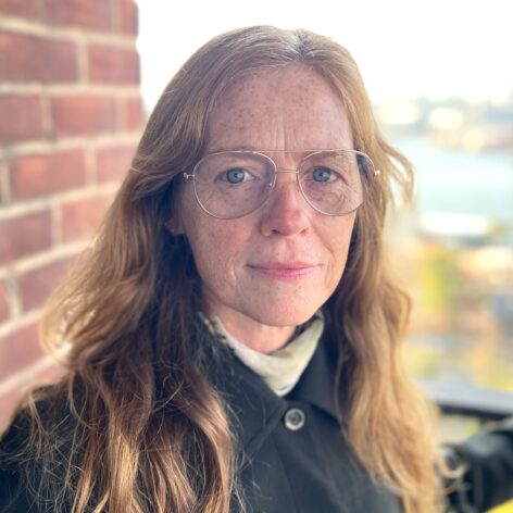 A white woman of middle age with long red hair and glasses, standing against a red brick background outdoors. Photo by Simon Sherber-Brennan.
