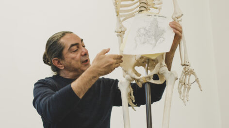 A photo of Jaime Ortega next to a human skeleton replica, holding up a picture of a pelvis. He is speaking and pointing towards the right side of the pelvis. Photo by Tori Lawrence.