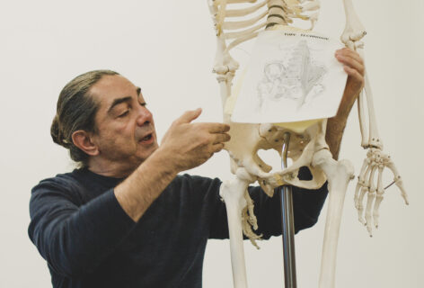 A photo of Jaime Ortega next to a human skeleton replica, holding up a picture of a pelvis. He is speaking and pointing towards the right side of the pelvis. Photo by Tori Lawrence.