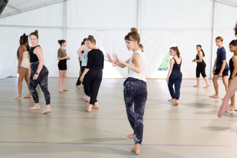 Photo of Joanna Kotze teaching dancers in a large studio. She has brown hair in a messy bun and bangs. She is demonstrating a movement with her hands bending at the elbow and palms facing away from her face, fingers spread. The students are behind her paying attention and adjusting. Photo by Jhsumedia.