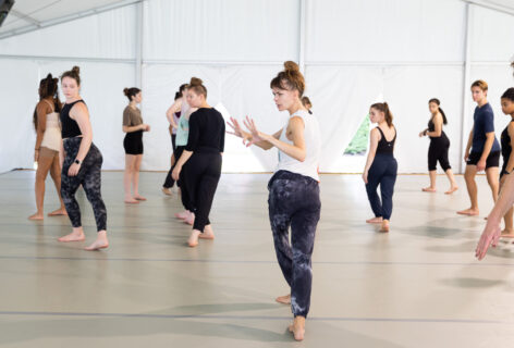 Photo of Joanna Kotze teaching dancers in a large studio. She has brown hair in a messy bun and bangs. She is demonstrating a movement with her hands bending at the elbow and palms facing away from her face, fingers spread. The students are behind her paying attention and adjusting. Photo by Jhsumedia.