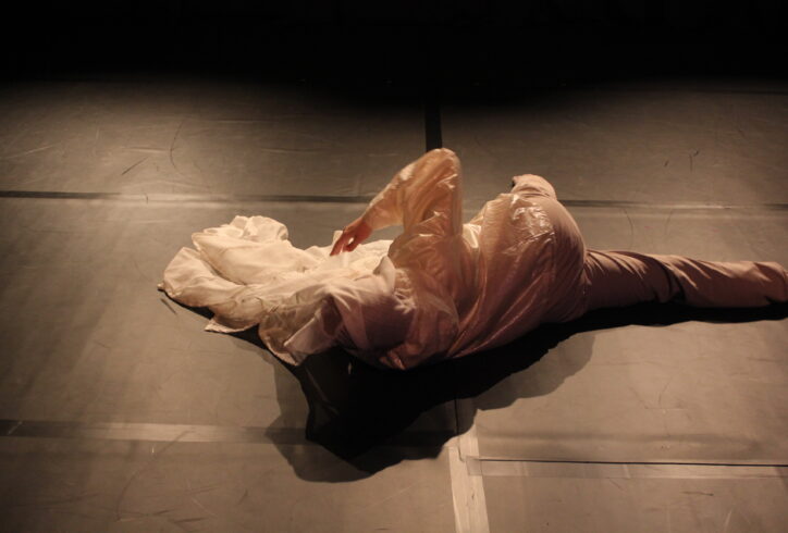 Kari rolls on the floor in beige athletic pants and an iridescent white jacket with sheer white fabric wrapped around their head and draped onto the floor. They are mid roll and their arm is in the air with their elbow bent. Photo courtesy of the artist.