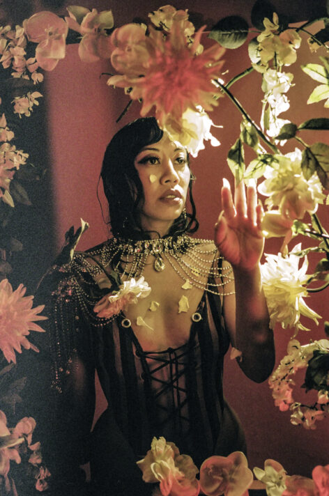 Portrait of Kat Sotelo. She is tan with slicked black hair and stands inside a deep red backdrop framed by white flowers. She is wearing a brown/black striped body suit, her shoulders adorned with jewels, as she caresses a dangling leaf with her uplifted left hand. Photo by Sarrah Danziger.