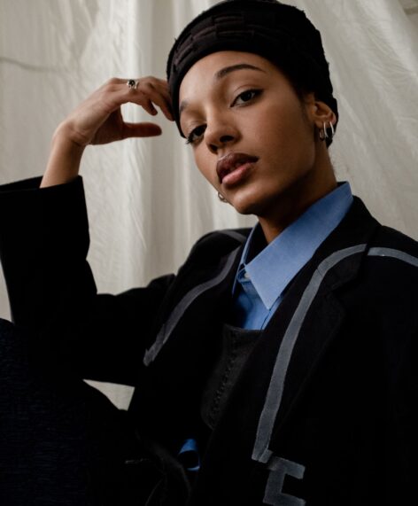 A photo of Katherine P. De La Cruz posing for Official Rebrand. She wears a black blazer with gray paint details and a blue button up. She rests her head on her arm propped up on her knee. She wears a black hat and looks into the camera. Photo by Jordi Perez.