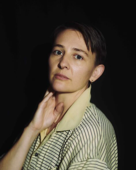Katy Pyle, a white genderqueer human, looks at the camera with a tilted head with their right hand by their neck in a relaxed gesture. Photo by Yael Malka.