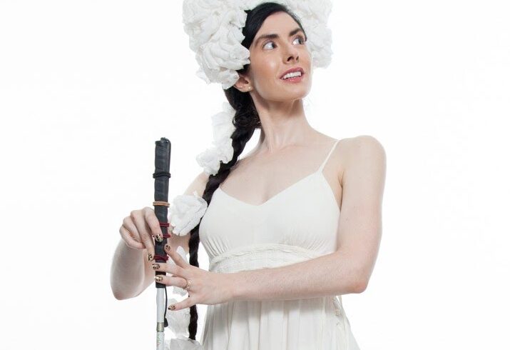 Krishna, a thin, pale skinned woman with long dark hair pulled into a braid, poses on a studio stage. She is wearing a simple white dress with her shoulders exposed and a fluffy white headdress perched on her forehead and wrapping down her braid. She looks over her left shoulder as she lightly holds a straight cane between her hands and points her left foot behind her. Photo Credit: Krishna Washburn & Micaela Mamede