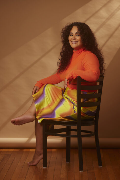 Portrait of Kristine sitting in a chair smiling at the camera. She has wavy hair and wears an orange long sleeve top and a colorful yellow skirt.Photo by Razzaq Manley.