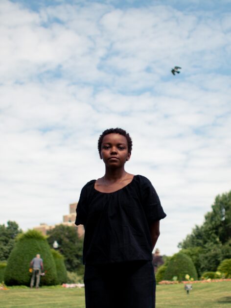 Laila stands in an open green field. She is a brown skinned Black woman with cropped short black hair. She wears a black blouse and gold chain. She stares serenely into the camera. Photo by Bailey Bailey.