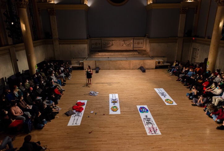Anabella Lenzu performing for MR@Judson. She stands speaking into a mic. Large illustration of skeletons are spread on the floor as well as other small objects. Audience members watch attentively. Photo by Rachel Keane.