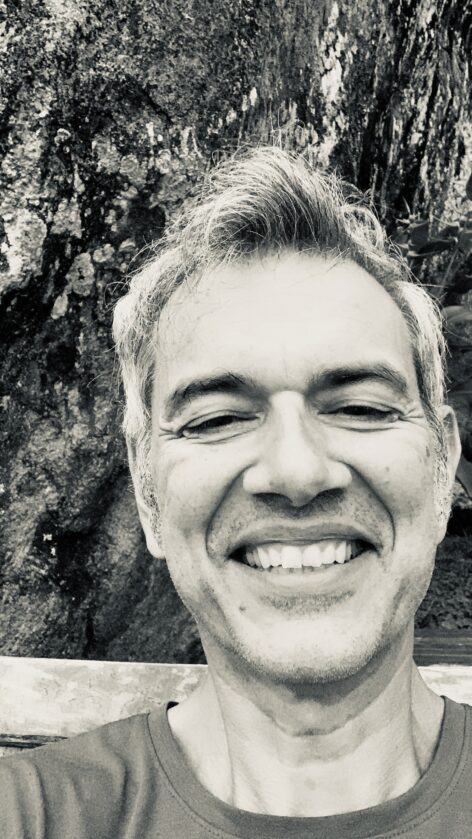 Frontal black and white headshot of a middle aged white male, salt and pepper hair, no facial hair, open smile, eyes half shut, against a rocky dark grey surface in the background. Photo/Image by Valentina Lepecki.