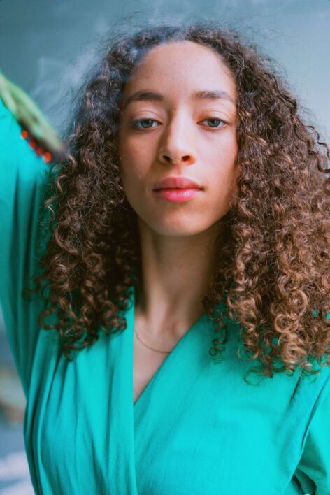 Rochelle Wilbun looks into the camera with a calm expression. She has curly light brown hair,  green eyes and a wears a bright green top. Photo courtesy of the artist.