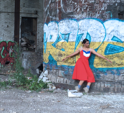 Nicole Soto Rodríguez dances in front of a wall with Graffiti murals. She wears a red dress and stands with her knees slightly bent. Her arm extend out from her torso. Photo courtesy of the artist.