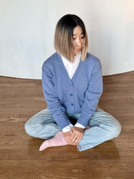 An Asian woman with blue jeans and a cardigan is sitting on the floor with crossed legs. Her body faces front, but her head faces left with her eyes softly looking down. Photo by Ron Nicolaysen.