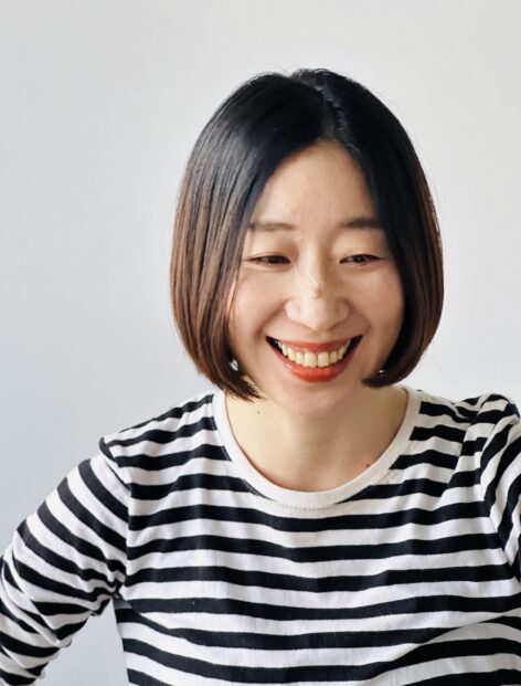 Photo of Maho smiling softly with white background. She has bob hair and wearing a striped shirt. Photo by Ron Nicolaysen.