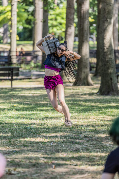 Photo of Stacy Lynn Smith performing BOOM WALK outside at the Williamsbridge Oval in the Bronx. A vintage boom box is balanced on one shoulder while their finger is in their mouth like a fish hook, causing them to spin in wide circles throughout the grass, long box braids swinging as they respond to the recorded cassette track that is BOOM WALK. Photo by Shintaro Ueyama.