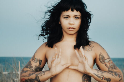 Photo of Marielys Burgos Meléndez a looking into the camera. She is topless with her hands placed in front of her chest, thumbs almost touching and fingers extending pushing the palms outwards. Her arms are tattooed and the ocean in behind her. Photo by Kyle Kesses.