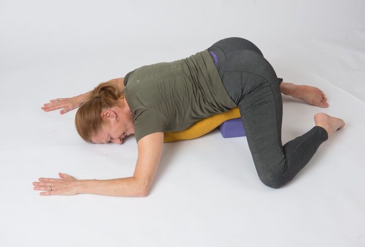 Martha Eddy demonstrating a Yoga prone position, Frog. A bolster and a block supporting her abdomen and chest. Face, forearm, and shins touching the ground. Photo by Serge Cashman.