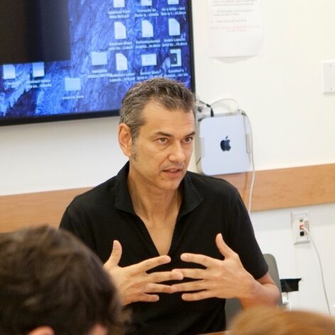 Andre wears a black shirt and sits in front of a screen. He gestures with both hands to his chest imd sentence. The tops of 2 heads in the foreground.