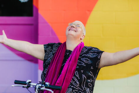 Petra Kuppers, a white queer disabled cis woman
of size with yellow glasses, shaved head, pink lipstick and a black
dotted top, smiles up to the sky, arms outstretched, embracing the
world. Her mobility scooter’s handlebar is visible at the bottom of the
image. She is in front of a multicolored wall: purple, pink, yellow and
orange. Photo by Tamara Wade